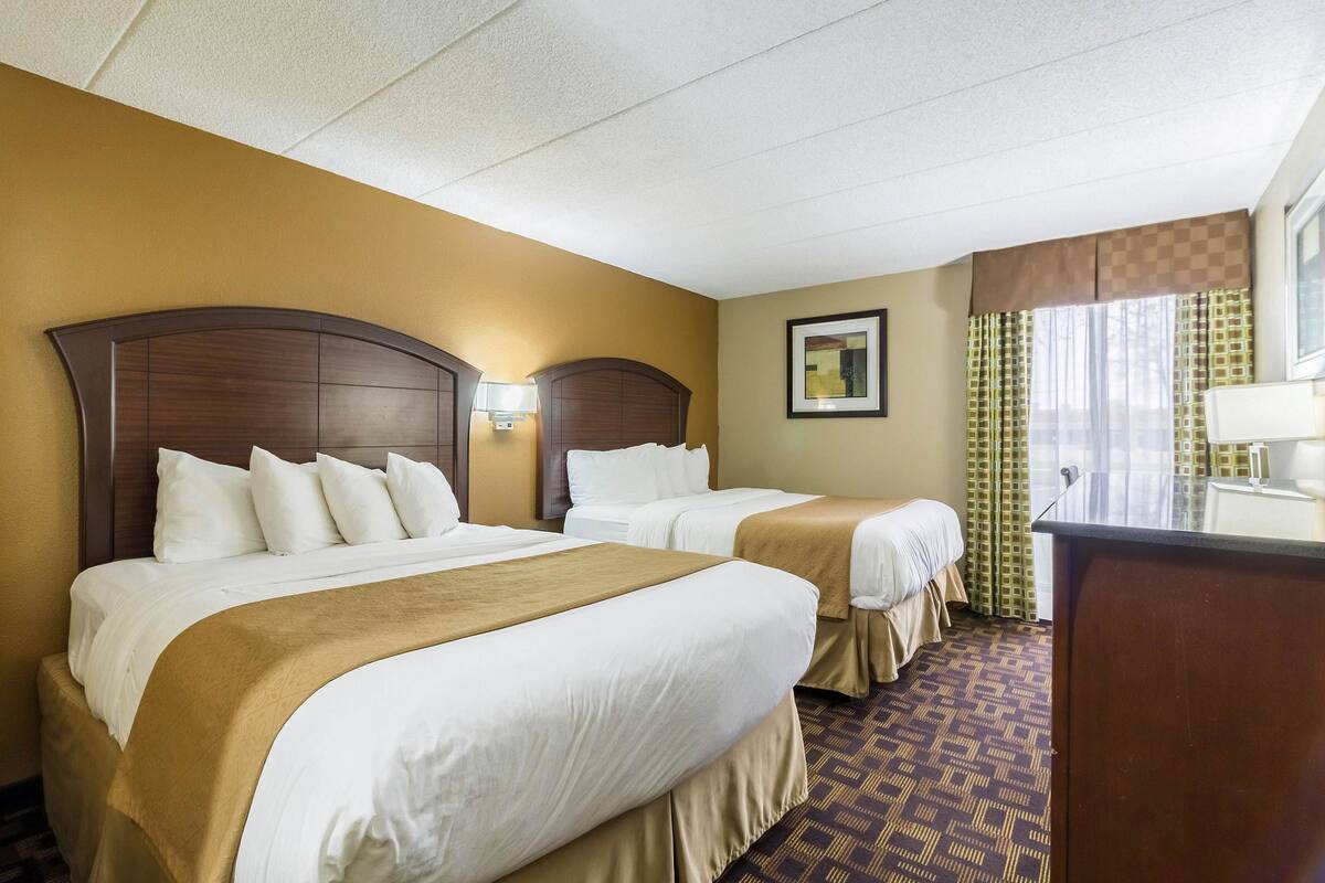 A hotel room with two beds, a desk, a window with curtains, and a framed picture on the wall.