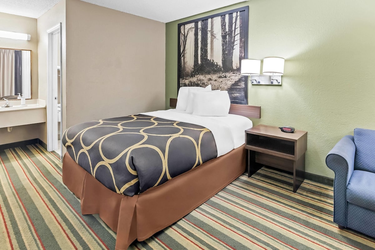A hotel room with a bed, a nightstand, a chair, a painting on the wall, and a bathroom visible through an open door.