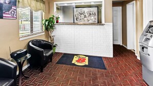Un área de recepción con pared de ladrillo blanco, mostrador de recepción, un sillón de cuero negro, una mesa de vidrio, alfombra roja y un letrero de Super 8 by Wyndham.