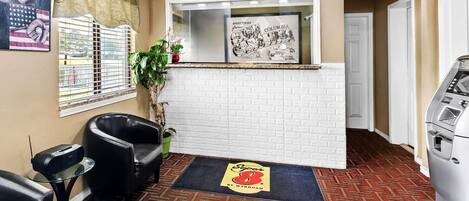 Un área de recepción con pared de ladrillo blanco, un mostrador de recepción, una radio Sony, dos sillones de cuero negro, una planta y el logotipo de Super 8 by Wyndham en la alfombra.