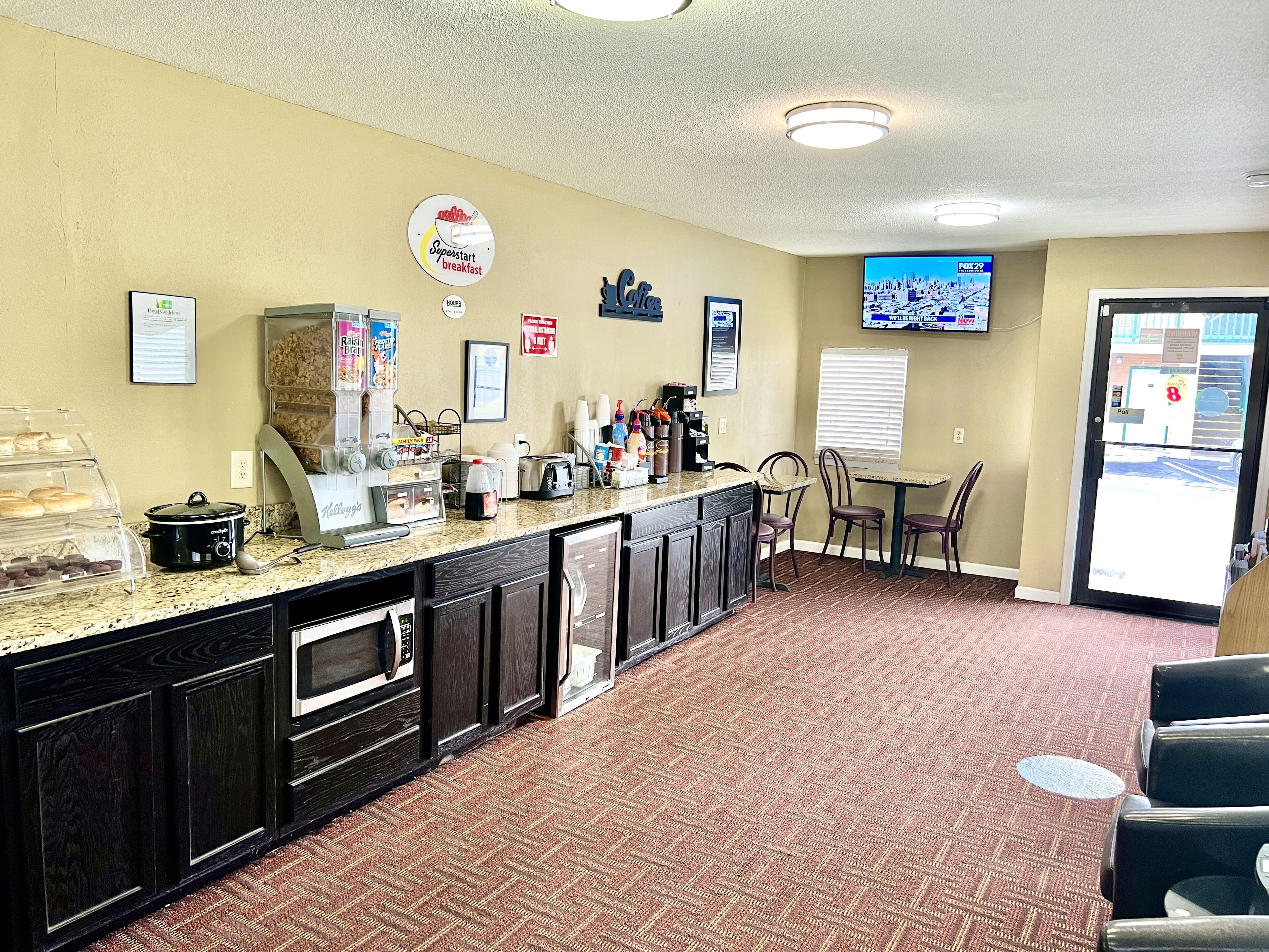 A hotel lobby with a breakfast bar, a coffee station, and a TV displaying a cityscape.