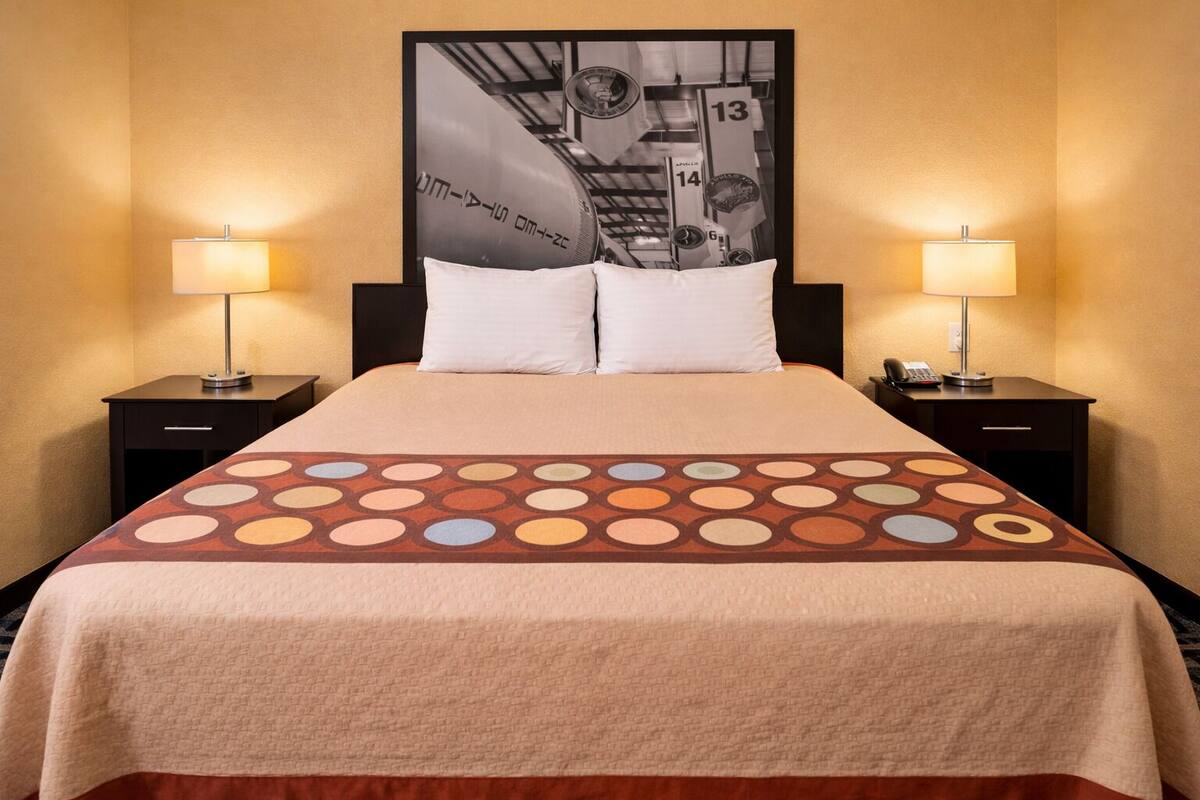 Quarto de hotel com uma cama, duas mesas de cabeceira com luminárias, um cartaz grande na parede e um edredom estampado.