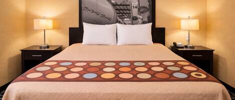 A hotel room with a bed, two bedside tables with lamps, a large poster on the wall, and a patterned bedspread.