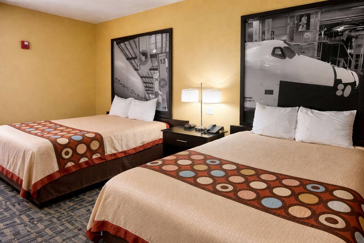 Quarto de hotel com duas camas, uma mesa de cabeceira, um abajur e uma janela grande com vista para um ônibus espacial.