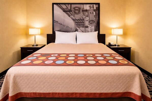 A hotel room with a large bed, two bedside tables with lamps, a headboard with a framed picture, and a patterned bedspread.