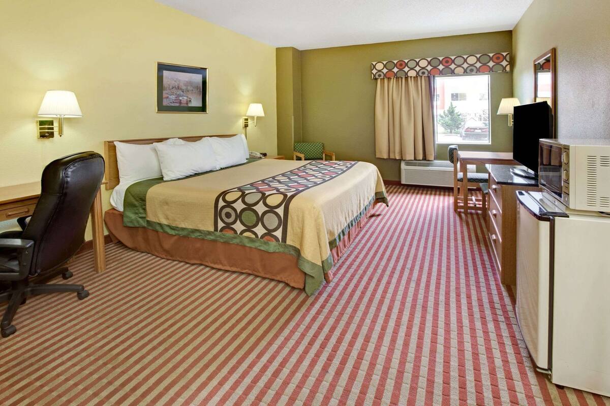 Quarto de hotel com cama, escrivaninha, cadeira, televisão e janela com cortinas.