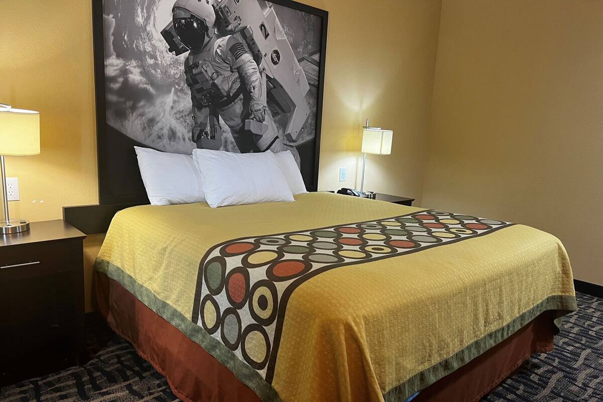 Quarto de hotel com uma cama grande, mesa de cabeceira com abajur, televisão na parede e piso coberto por carpete.