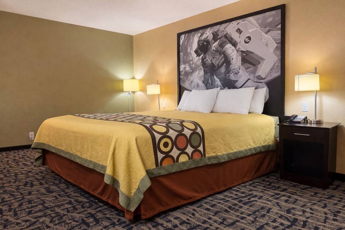 Quarto de hotel com cama, duas luminárias de cabeceira, mesa de cabeceira com telefone e um quadro grande na parede.