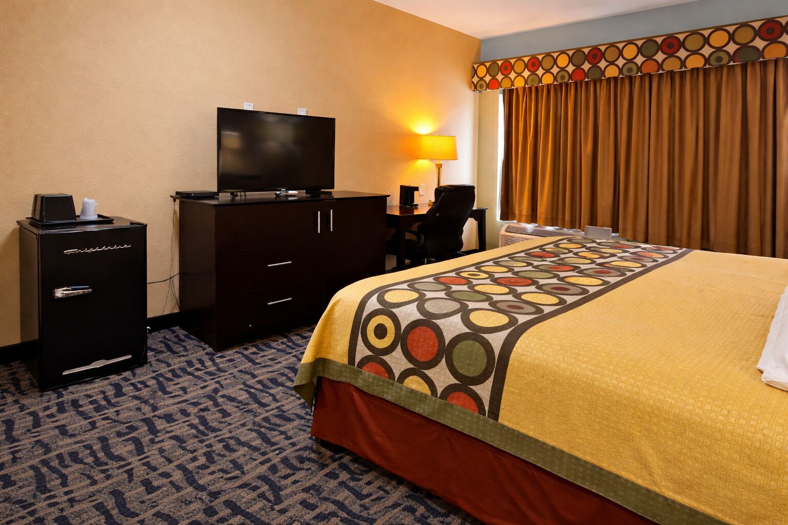 A hotel room with a bed, a TV, a desk, a chair, a minibar, and a patterned carpet.