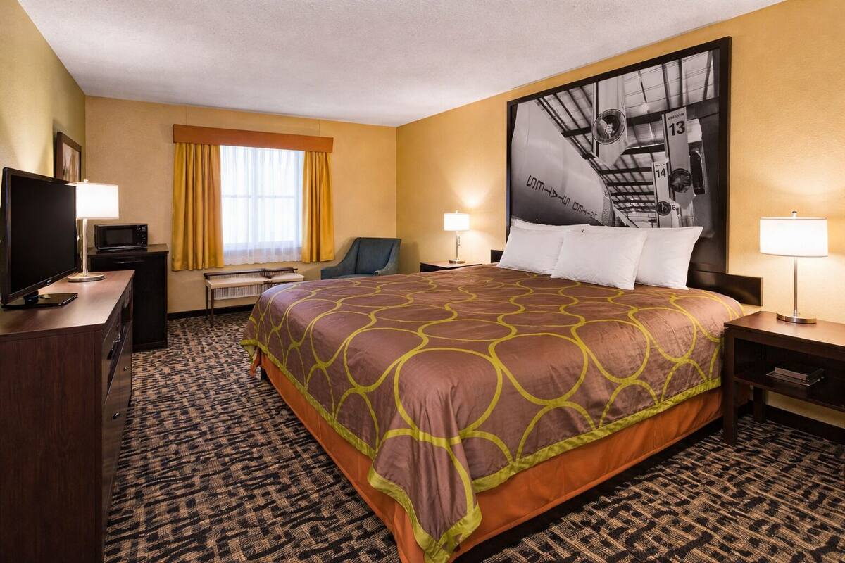 Quarto de hotel com uma cama grande, uma mesa de trabalho com cadeira, televisão e uma obra de arte na parede.