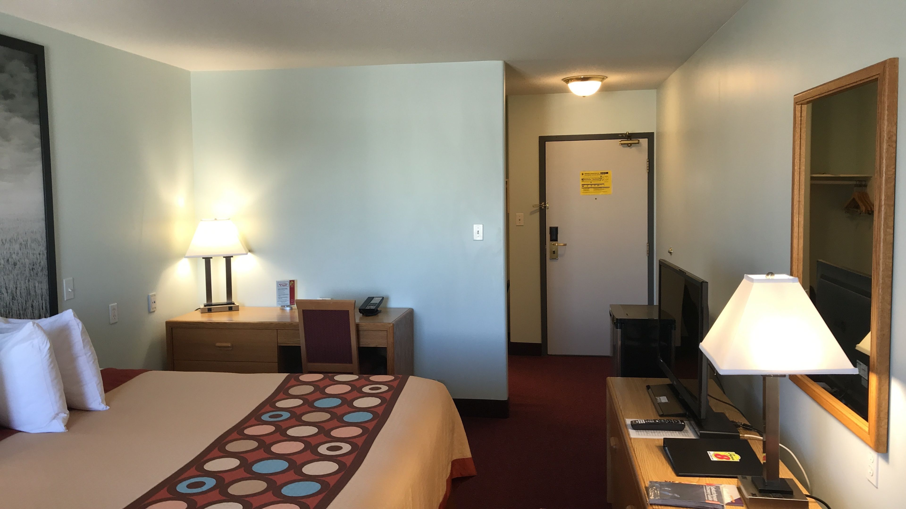 Habitación de hotel con cama, escritorio con lámpara, televisor y espejo.