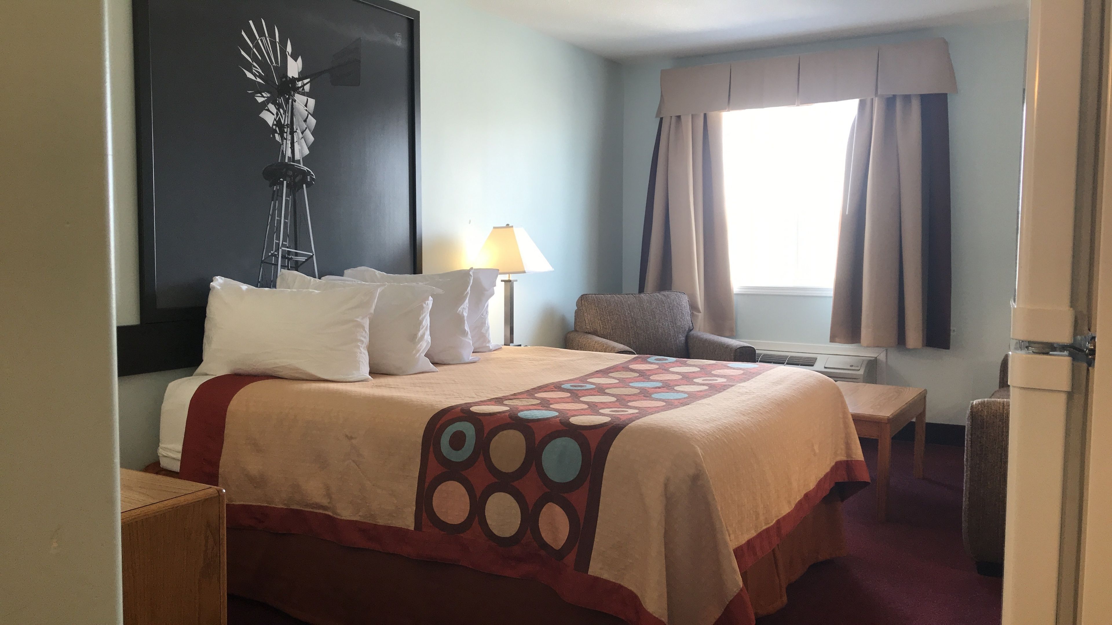Una habitación de hotel con cama, una silla, escritorio, lámpara y ventana con cortinas.