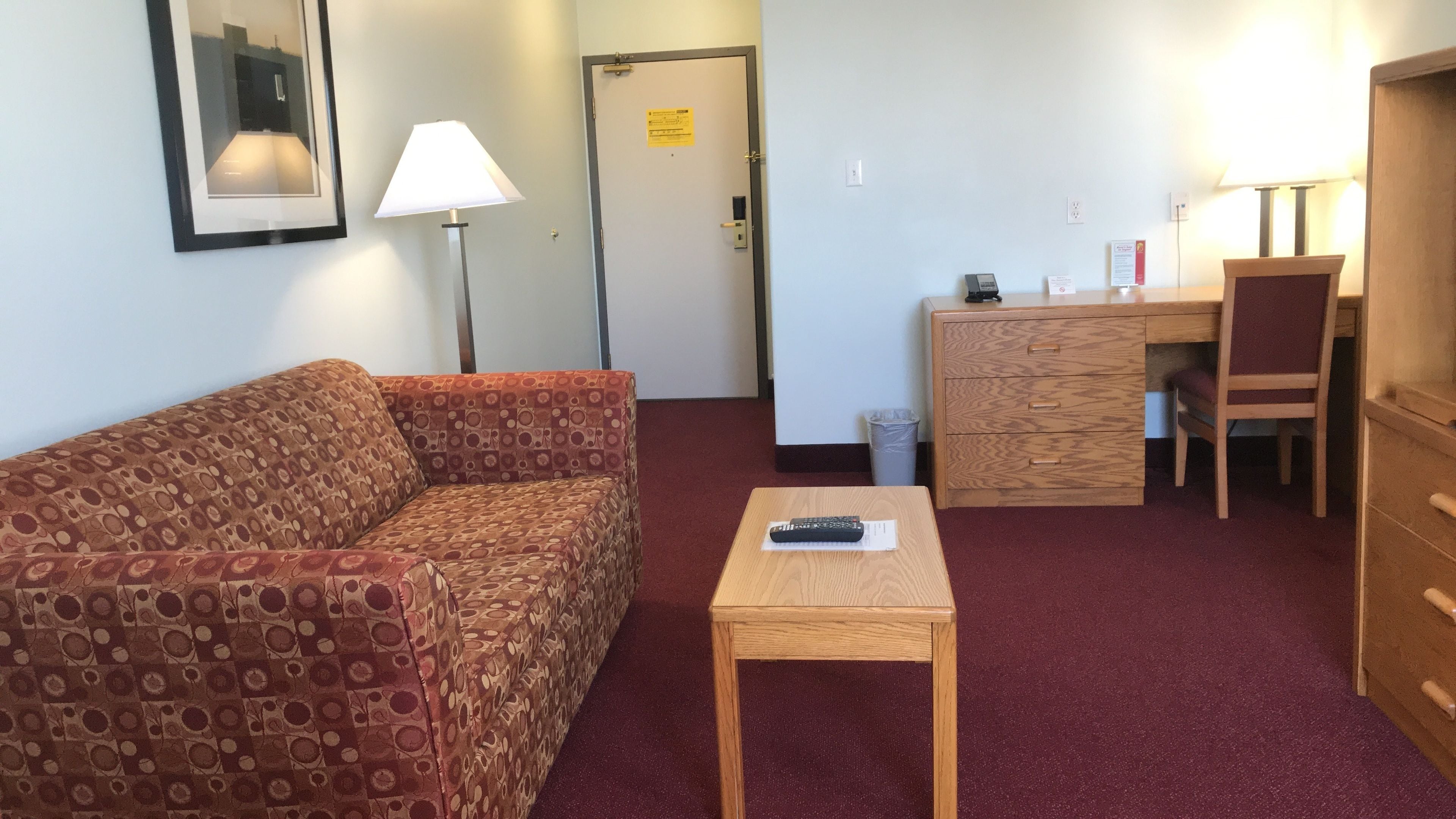 A furnished room with a sofa, a small table, a desk with a chair, a lamp, and a door with a sign.