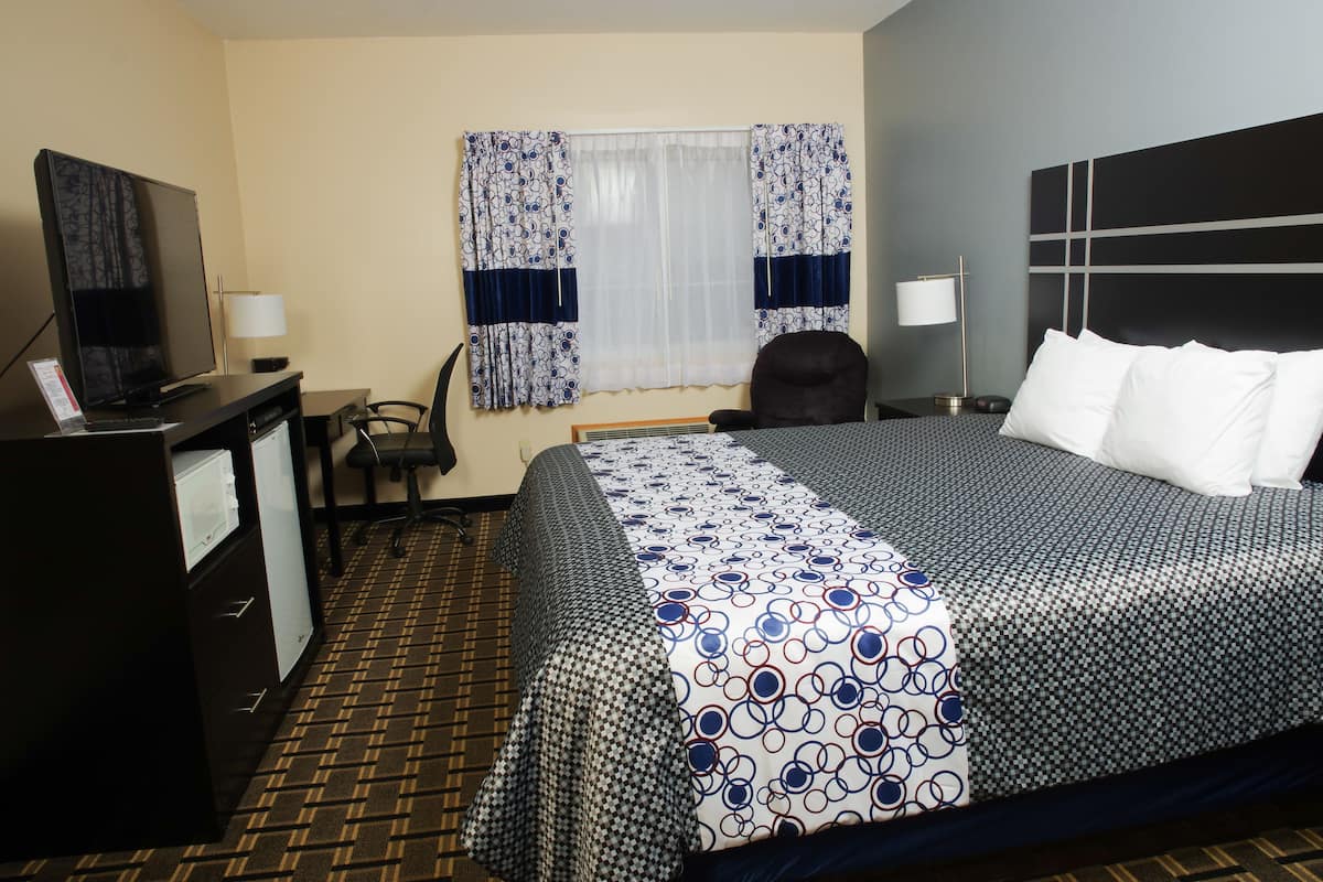 A hotel room with a bed, a desk with a television, a chair, a window with curtains, and a nightstand with a lamp.