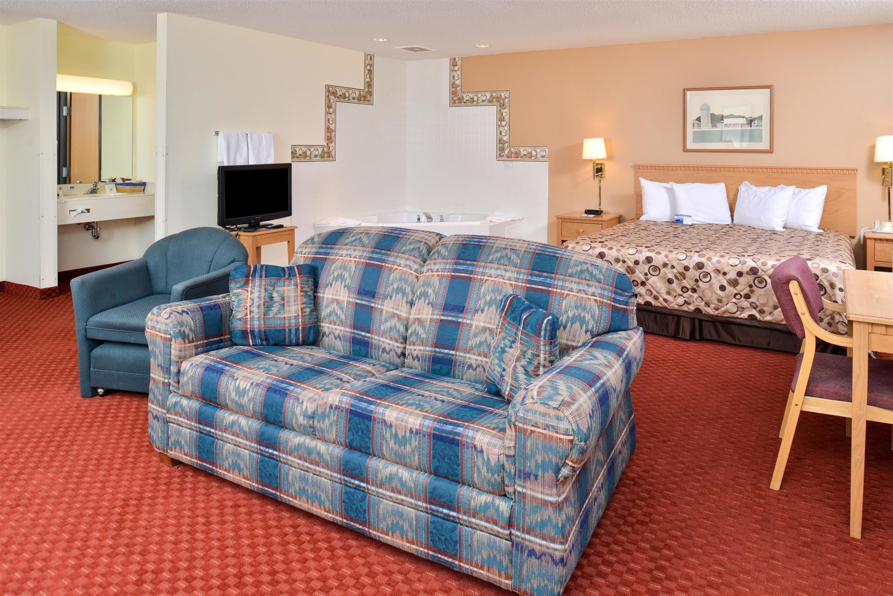 A hotel room with a plaid sofa, a bed with patterned bedding, a desk with a chair, a television, and a bathroom with a mirror and sink.