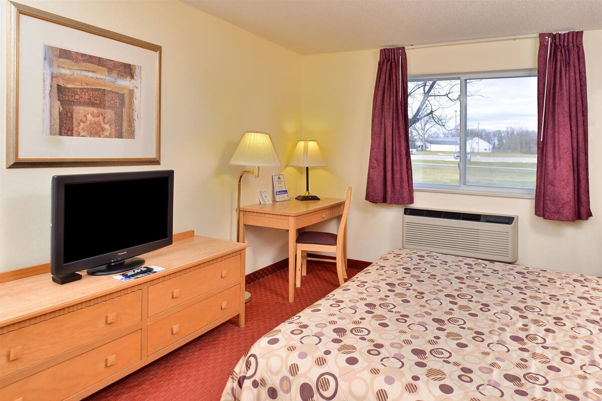 A hotel room with a bed, a desk with a chair, a television, a framed picture, and a window with curtains.