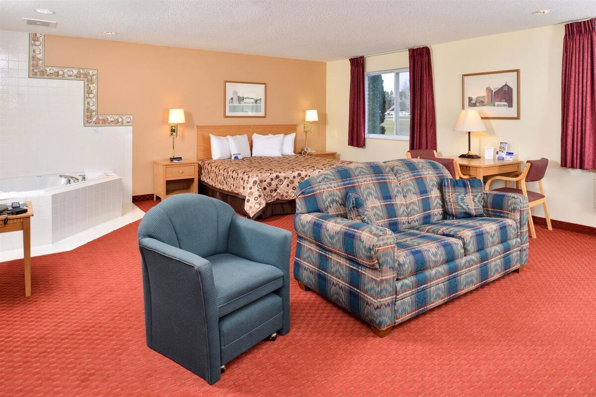 A hotel room with a bed, a sofa, a chair, a desk, a lamp, a window with curtains, a painting, and a bathtub.