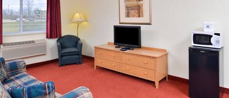 A hotel room with a red carpet, a sofa, a TV on a wooden dresser, a microwave, and a window with a view of a parking lot.