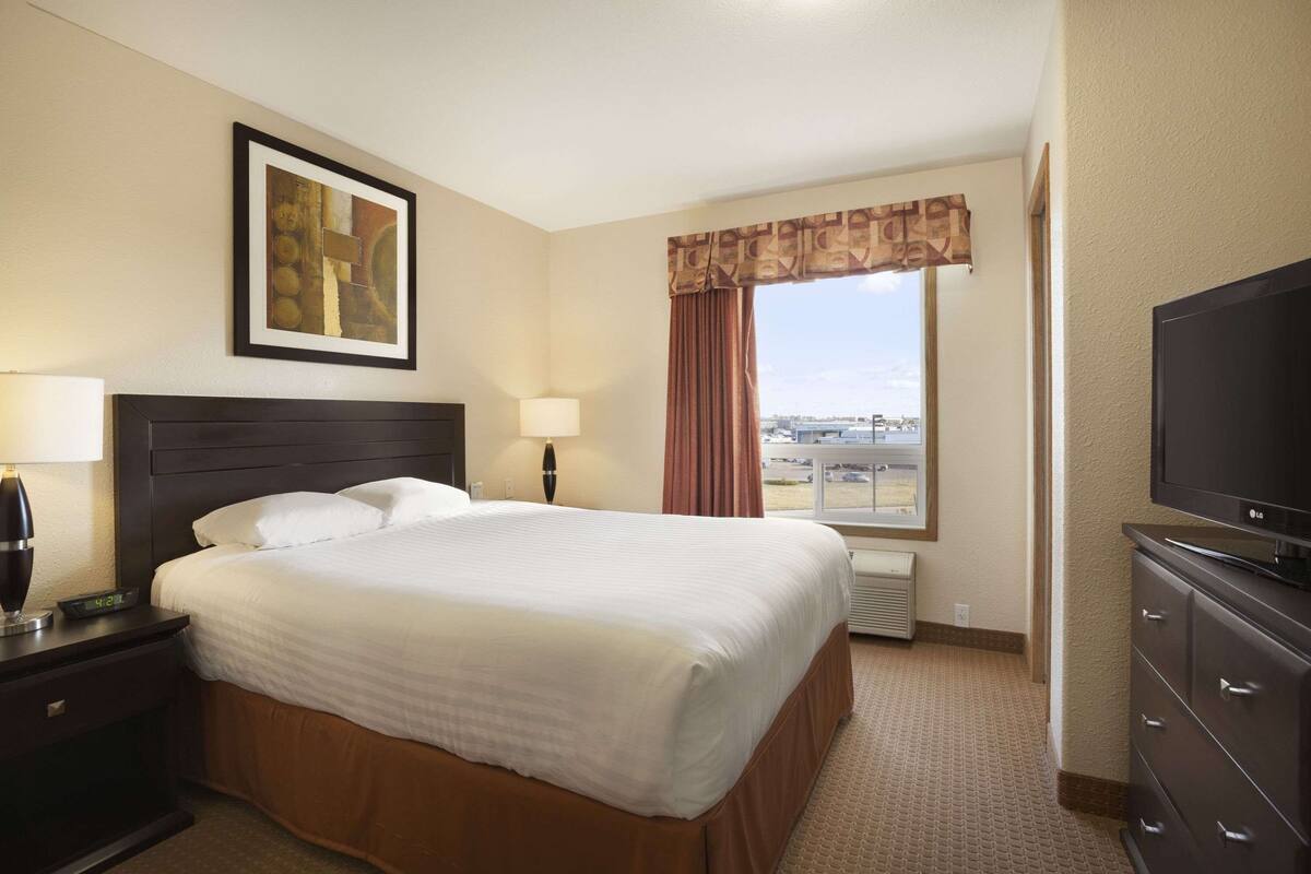 Habitación de hotel con una cama grande, un televisor, un escritorio con lámpara, un cuadro en la pared y una ventana con cortinas.