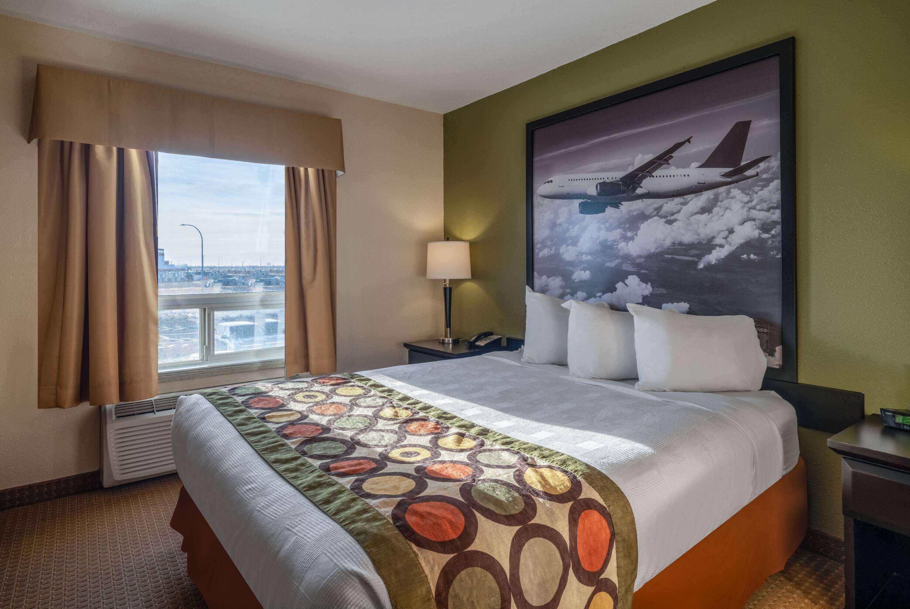 Une chambre d’hôtel avec un grand lit, une fenêtre avec des rideaux, une table de chevet, une lampe et un tableau représentant un avion.