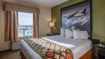 Une chambre d’hôtel avec un grand lit, une fenêtre avec des rideaux, une table de chevet, une lampe et un tableau représentant un avion.