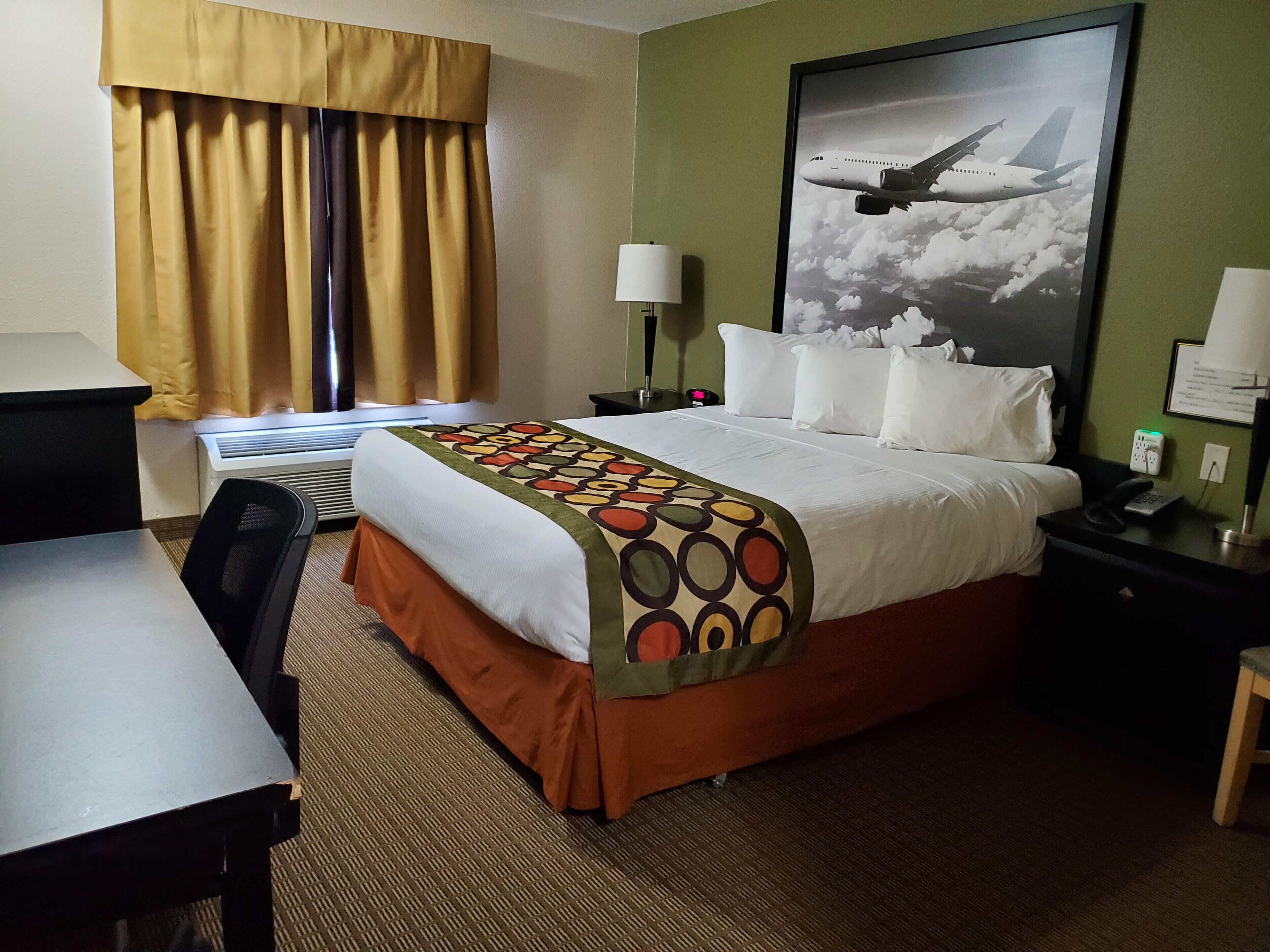 Habitación de hotel con cama, escritorio, silla, lámpara, mesita de noche y un cuadro colgado en la pared de un avión.