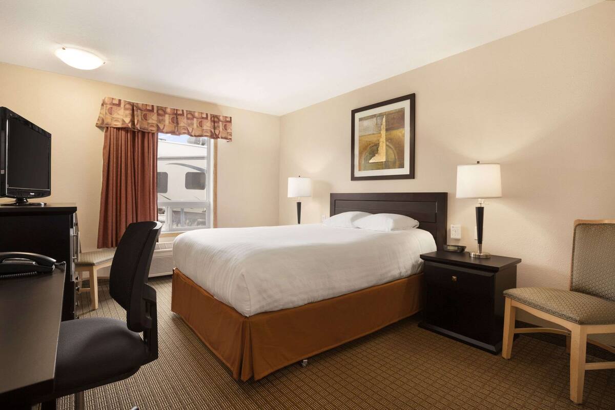 Una habitación de hotel con cama, escritorio con computadora, silla, mesita de noche con lámpara y un cuadro en la pared.