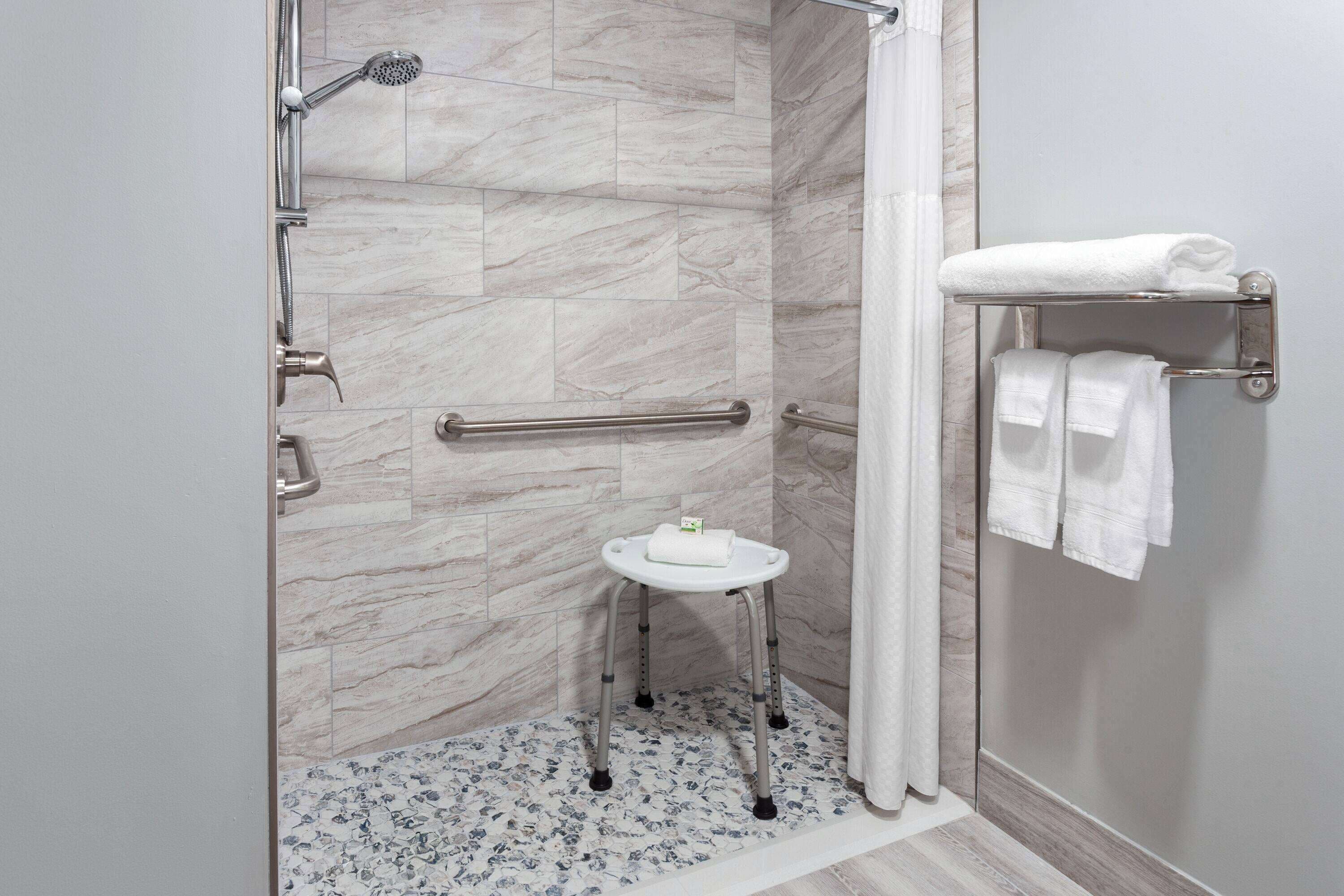 A shower area with a handrail, a small stool with a towel, and a shelf with towels.