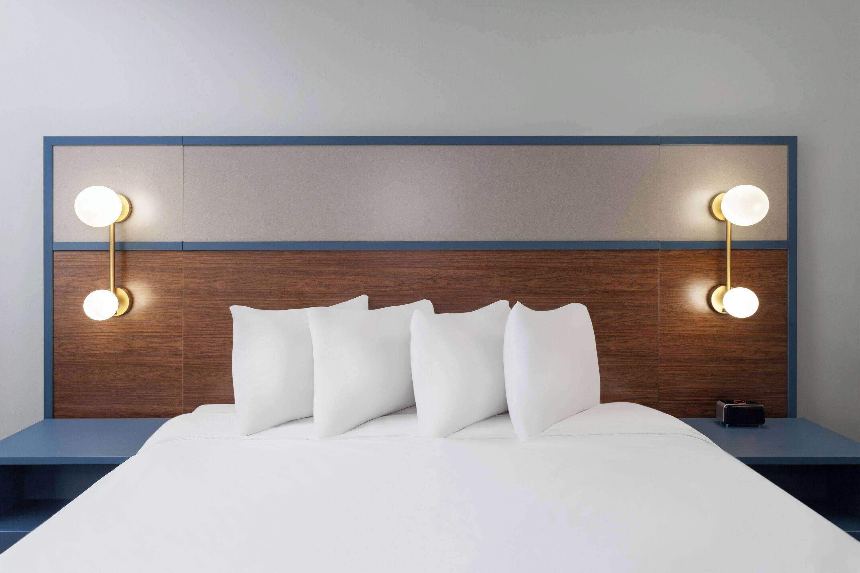 Una cama bien hecha con sábanas y almohadas blancas, un cabecero de madera con detalles azules y dos lámparas de noche con luces redondas.