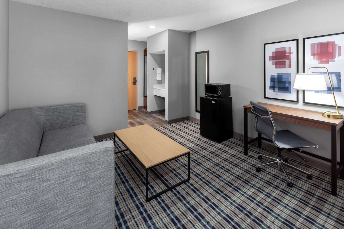 Een hotelkamer met een grijze bank, een houten tafel, een bureau met een stoel, een zwart tv-meubel en twee wanddecoraties met abstracte schilderijen.