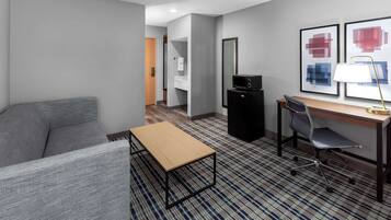 Une chambre d’hôtel avec un canapé gris, une table en bois, un bureau avec une chaise, un meuble TV noir et deux tableaux abstraits encadrés accrochés au mur.