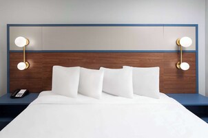 Una cama bien hecha con sábanas y almohadas blancas, un cabecero de madera con dos lámparas fijadas a la pared, y una mesita de noche con un teléfono y una lámpara.