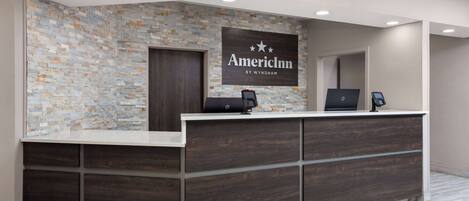 Een receptiebalie met een achtergrond van een stenen muur, een bord met daarop 'AmericInn by Wyndham' en twee laptops op de balie.