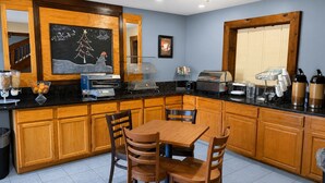 A kitchen with wooden cabinets, a dining table with chairs, a coffee station, and a chalkboard with holiday decorations. - Coratel Inn & Suites by Jasper Black River Falls (Black River Falls)