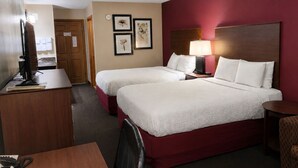 A hotel room with two beds, a desk with a computer, a chair, a lamp, and framed pictures on the wall.