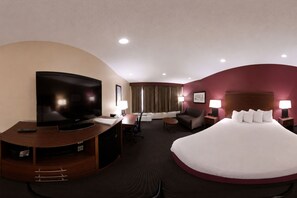 Ein Hotelzimmer mit einem großen Bett, einem Schreibtisch mit Fernseher, einem Sessel und einem Nachttisch mit Lampe.