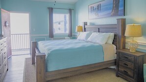 A bedroom with a bed, two bedside tables with lamps, a dresser, a window with curtains, and a painting on the wall. - Sea And Breeze Hotel And Condo (Tybee Island)