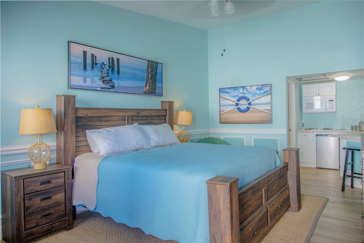 A bedroom with a bed, nightstands, a desk, and a kitchenette.