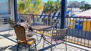 A balcony with outdoor seating, including chairs and a table, overlooking a street with buildings and palm trees. - Sea And Breeze Hotel And Condo (Tybee Island)