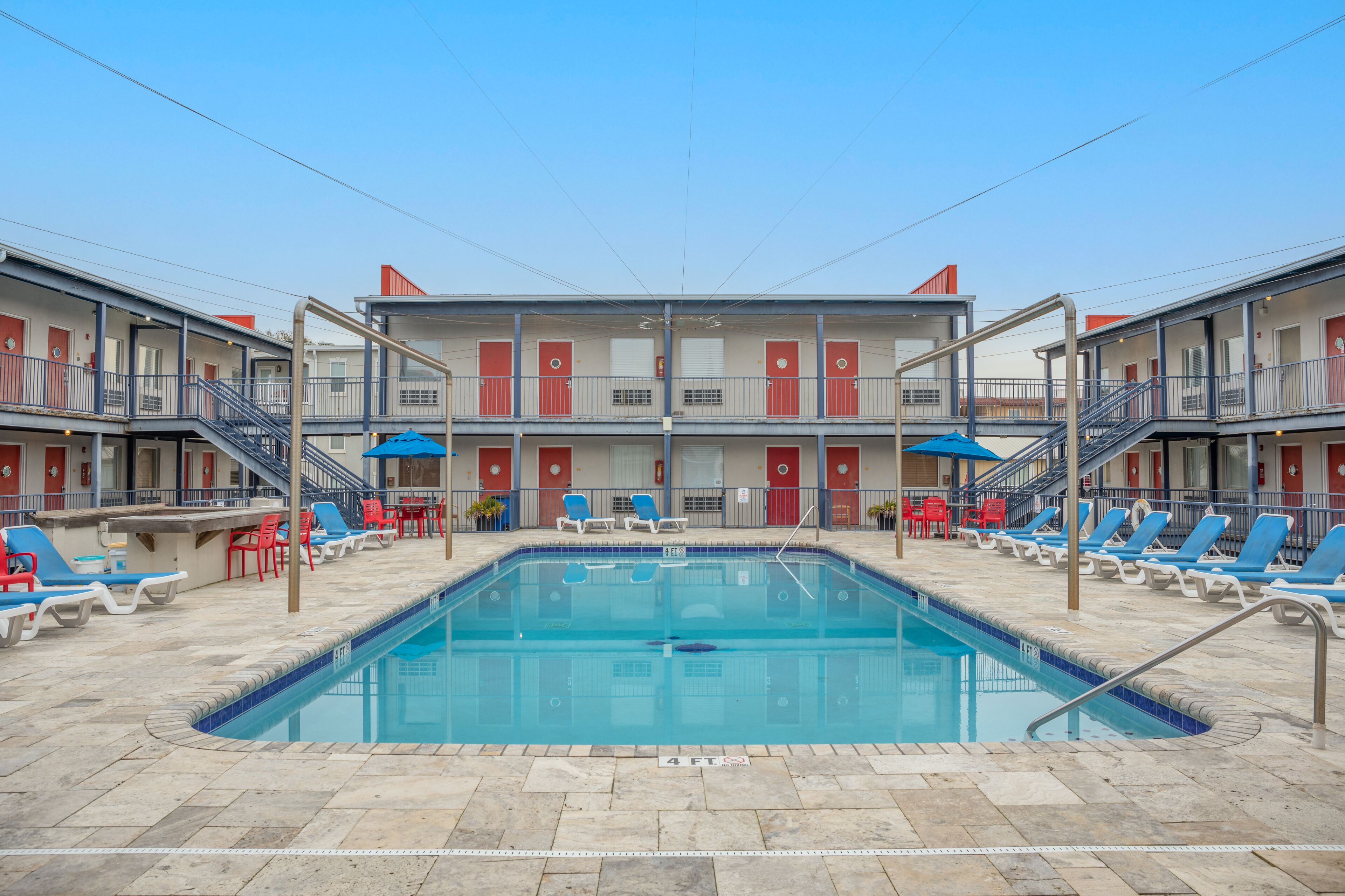 Un espace piscine doté de chaises longues et d’une piscine, le tout entouré d’un motel à deux étages, agrémenté de balcons et de portes rouges.