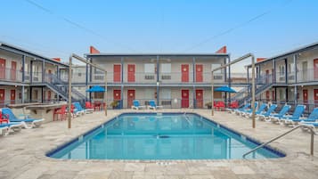 A pool area with lounge chairs and a swimming pool, surrounded by a two-story motel building with balconies and red doors.