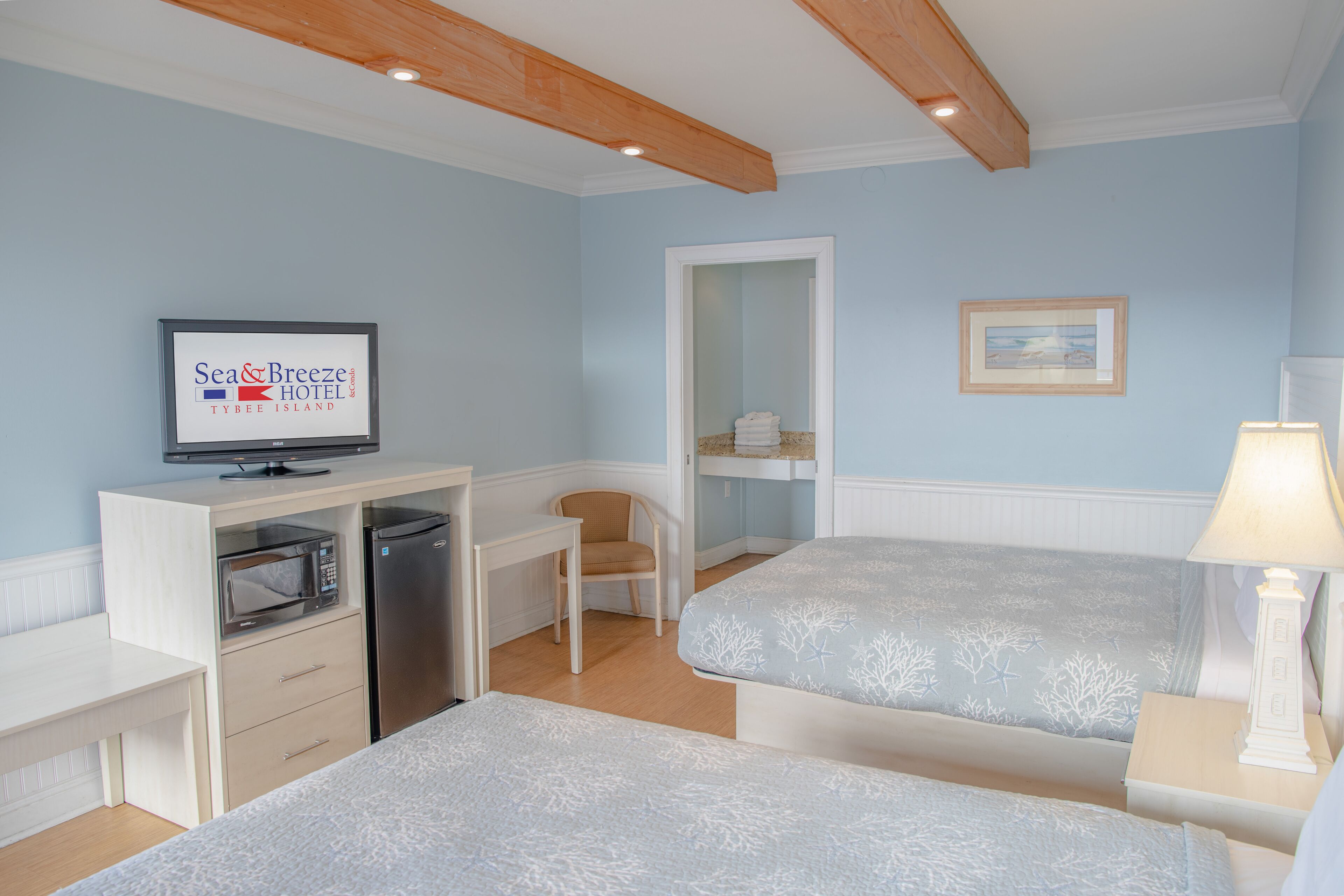 A hotel room with a television displaying the hotel name, a microwave, a refrigerator, two beds, a chair, a nightstand with a lamp, and a framed picture on the wall.