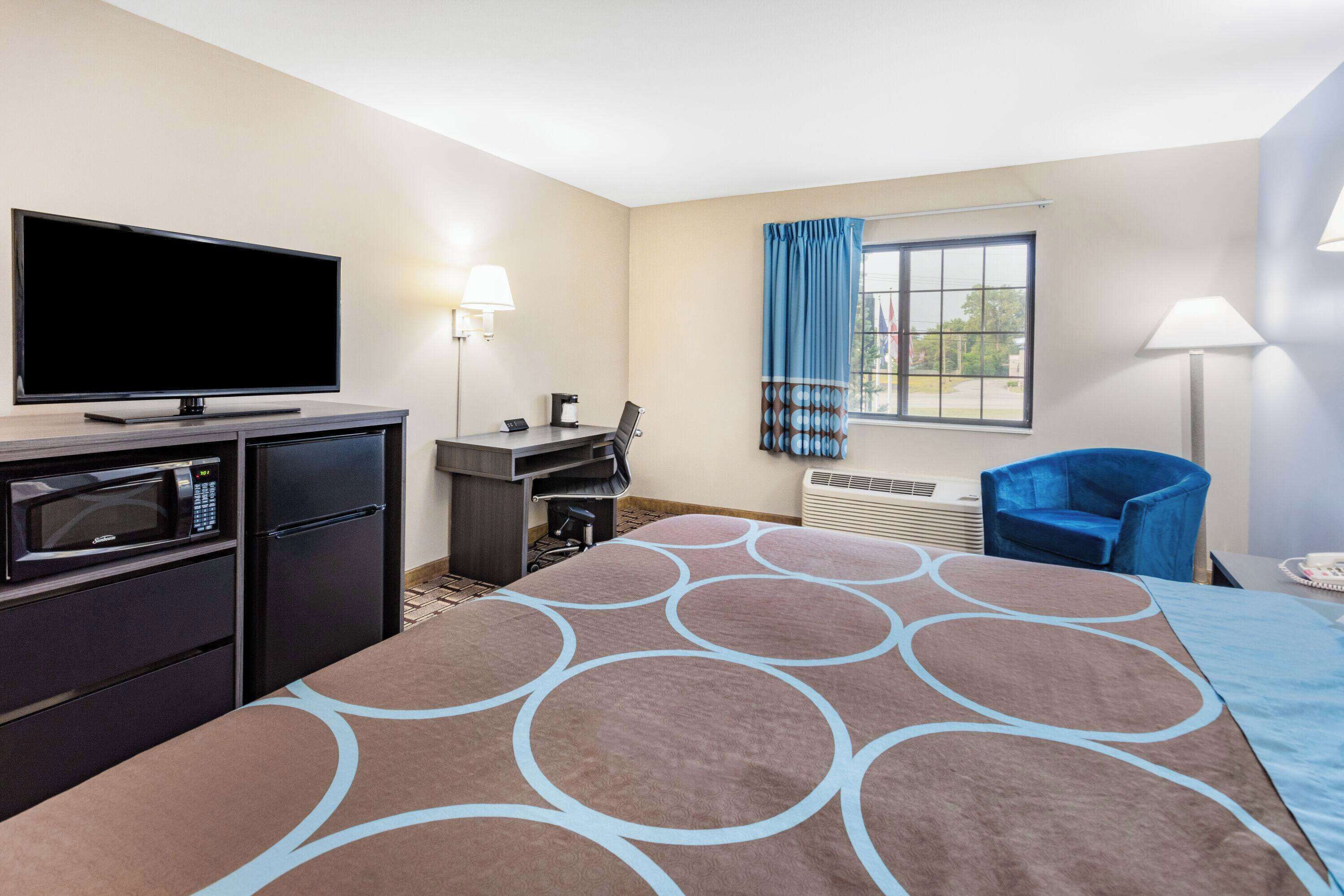 A hotel room with a flat-screen TV, a microwave, a refrigerator, a desk with a chair, a large bed with a patterned bedspread, a window with curtains, and a standing lamp.