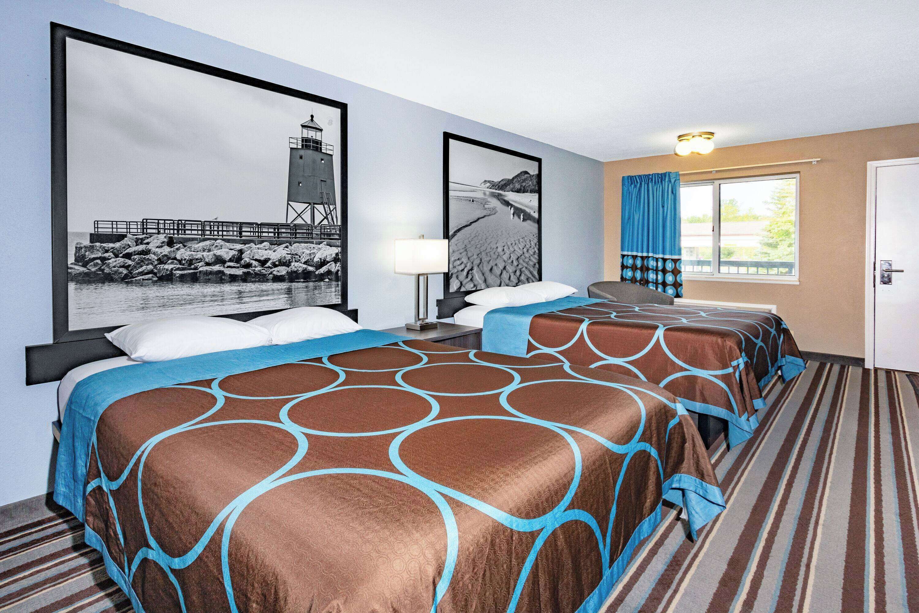 A hotel room with two beds, a large framed picture of a lighthouse, a window with curtains, and a striped carpet.