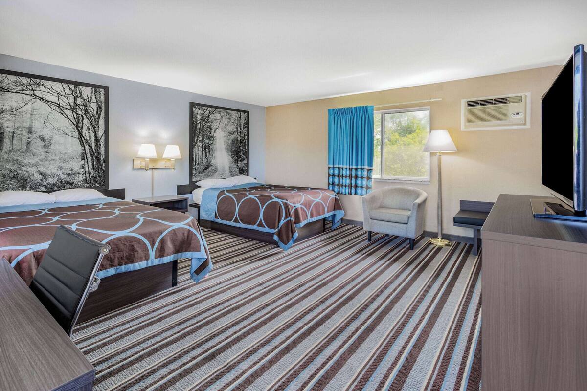 Habitación de hotel con dos camas, un escritorio, una silla, un ventanal con cortinas y obras de arte en las paredes.