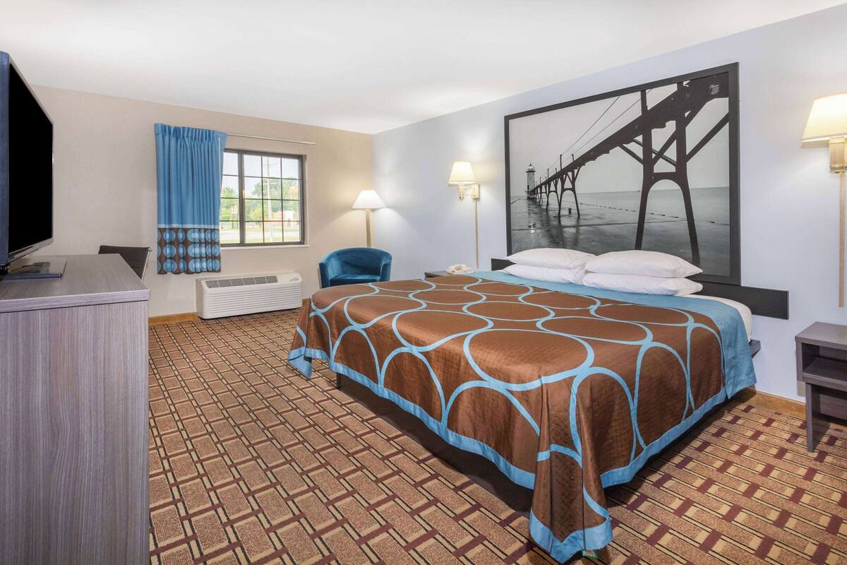 Habitación de hotel con cama, escritorio, silla, televisión y un amplio ventanal con cortinas.