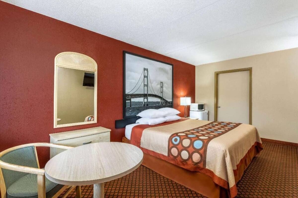 Habitación de hotel con cama, una mesa redonda, una silla, un espejo, un cuadro de un puente y una puerta.