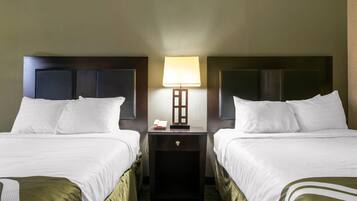 Una habitación de hotel con dos camas, una mesita de noche con lámpara y un cajón pequeño.