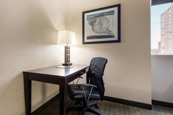A hotel room with a desk, chair, lamp, and a framed artwork on the wall.