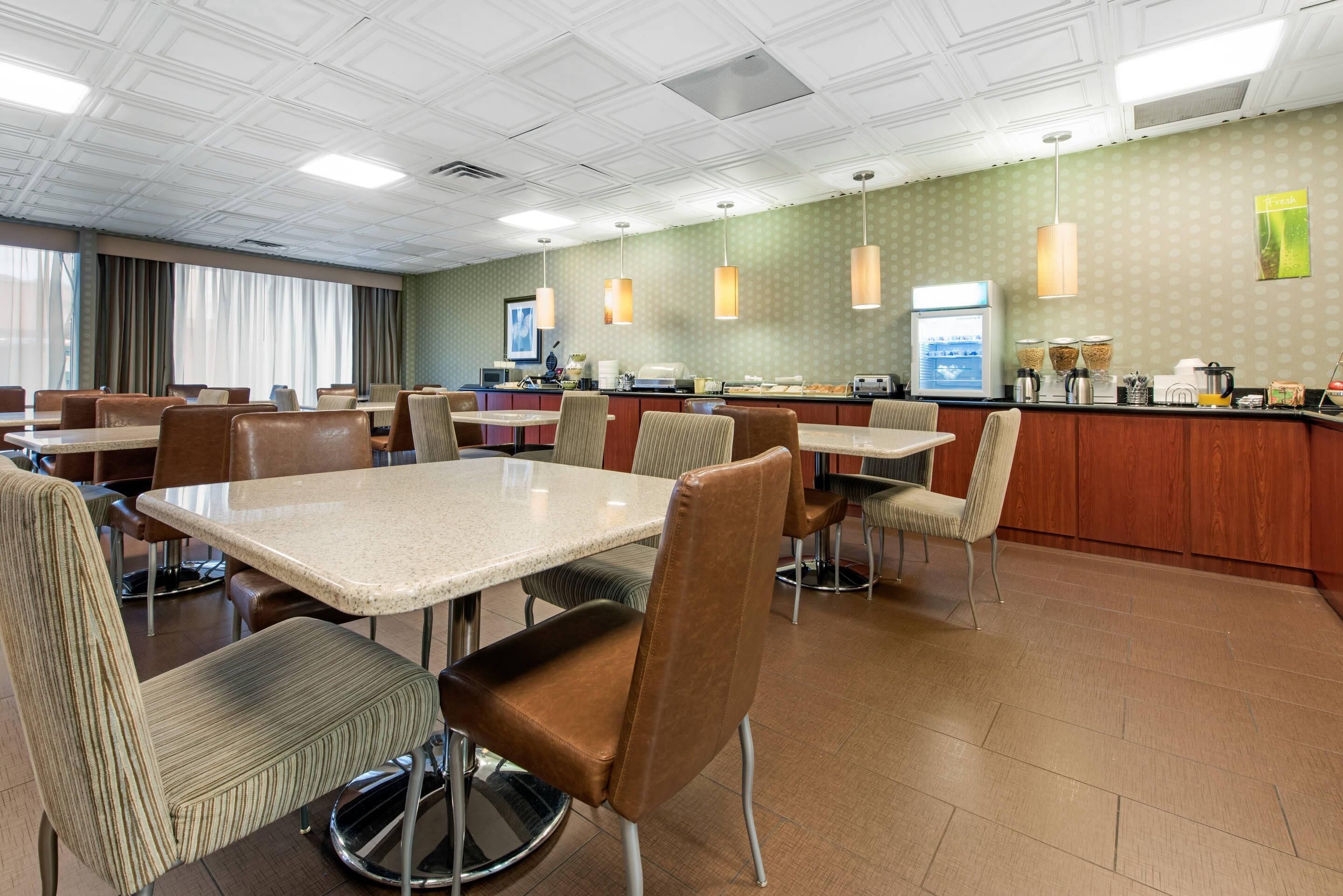 A hotel dining area with multiple tables and chairs, a counter with a coffee machine, and a wall with a 'Fresh' sign.